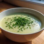 A close-up of a creamy, pale green bowl of leek soup topped generously with fresh chopped chives and black pepper.