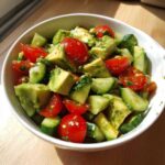 Close-up of a vibrant avocado salad featuring diced avocado, halved cherry tomatoes, and sliced cucumber tossed in a light dressing.