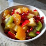 Close-up of a vibrant winter fruit salad featuring kiwi, orange segments, apple chunks, and bright red pomegranate seeds.