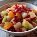 Close-up of a vibrant winter fruit salad featuring kiwi, orange segments, apple chunks, and bright pomegranate seeds.