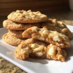 A stack of freshly baked white chocolate macadamia nut cookies, with one cookie broken in half showing a soft, chewy interior.