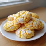 A stack of bright yellow lemon crinkle cookies dusted heavily with powdered sugar on a white plate.