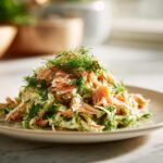 A mound of creamy salmon salad mixed with shredded vegetables and heavily garnished with fresh dill on a light plate.