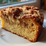 A close-up of a moist slice of apple crumb cake featuring visible apple chunks and a thick, golden brown crumb topping.