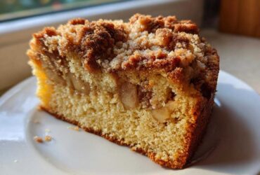 A close-up of a moist slice of apple crumb cake featuring visible apple chunks and a thick, golden brown crumb topping.