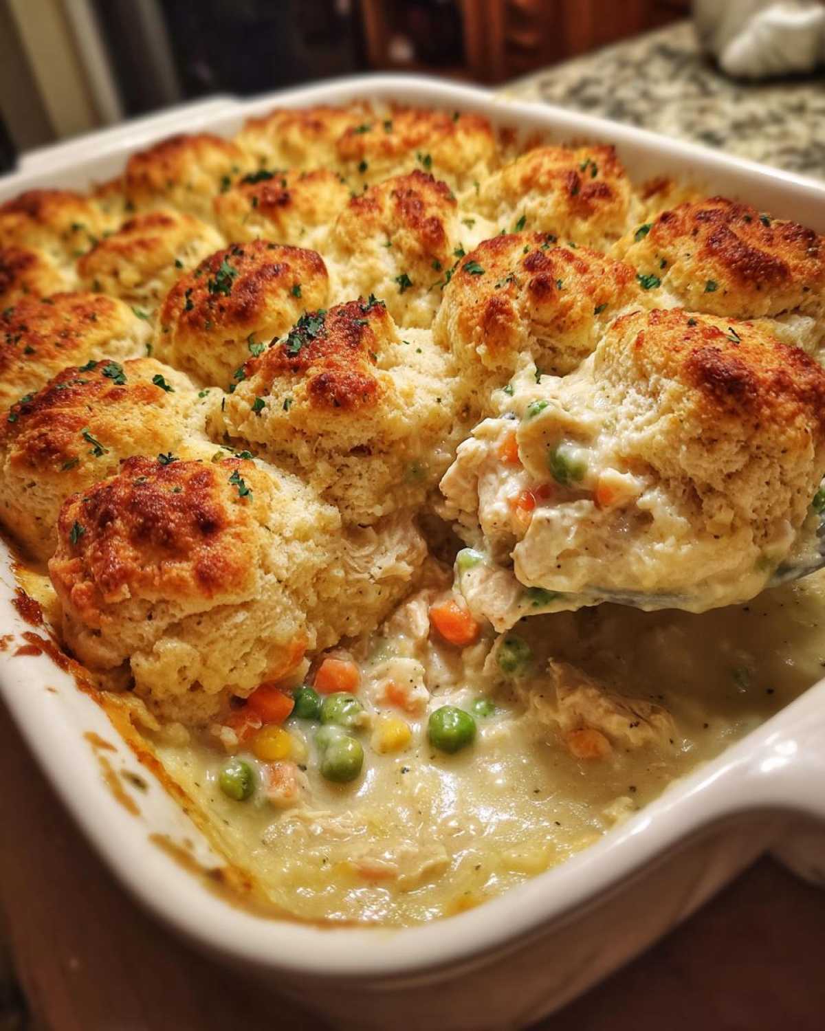 A spoonful of creamy chicken pot pie casserole filling being lifted from a baking dish topped with golden biscuits.