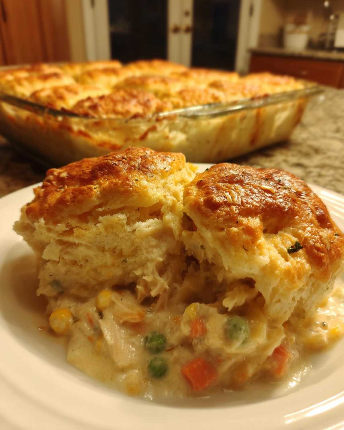 A serving of creamy chicken pot pie casserole topped with flaky biscuits, with the full dish in the background.