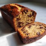 A close-up of a moist chocolate chip vegan banana bread loaf with one slice cut and resting in front.