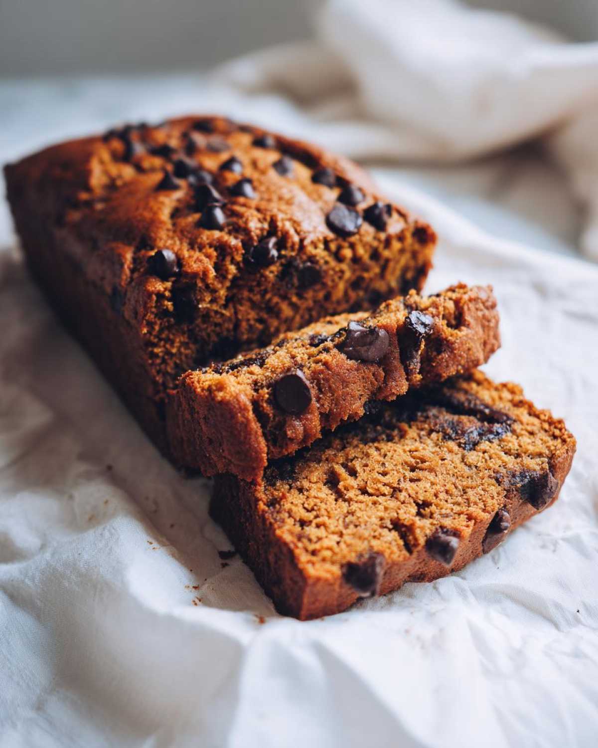 A close-up of a moist chocolate chip vegan banana bread loaf with two slices cut and leaning against it.