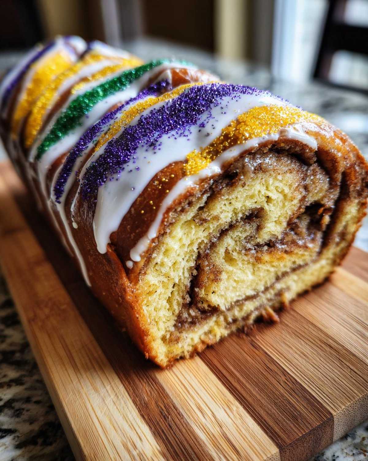A close-up cross-section of a homemade king cake loaf showing cinnamon swirl filling, topped with white icing and purple, green, and gold sprinkles.