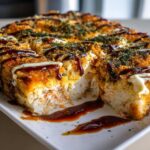 A close-up, mouth-watering view of a baked sushi bake, cut into squares, drizzled with dark sauce and mayo.