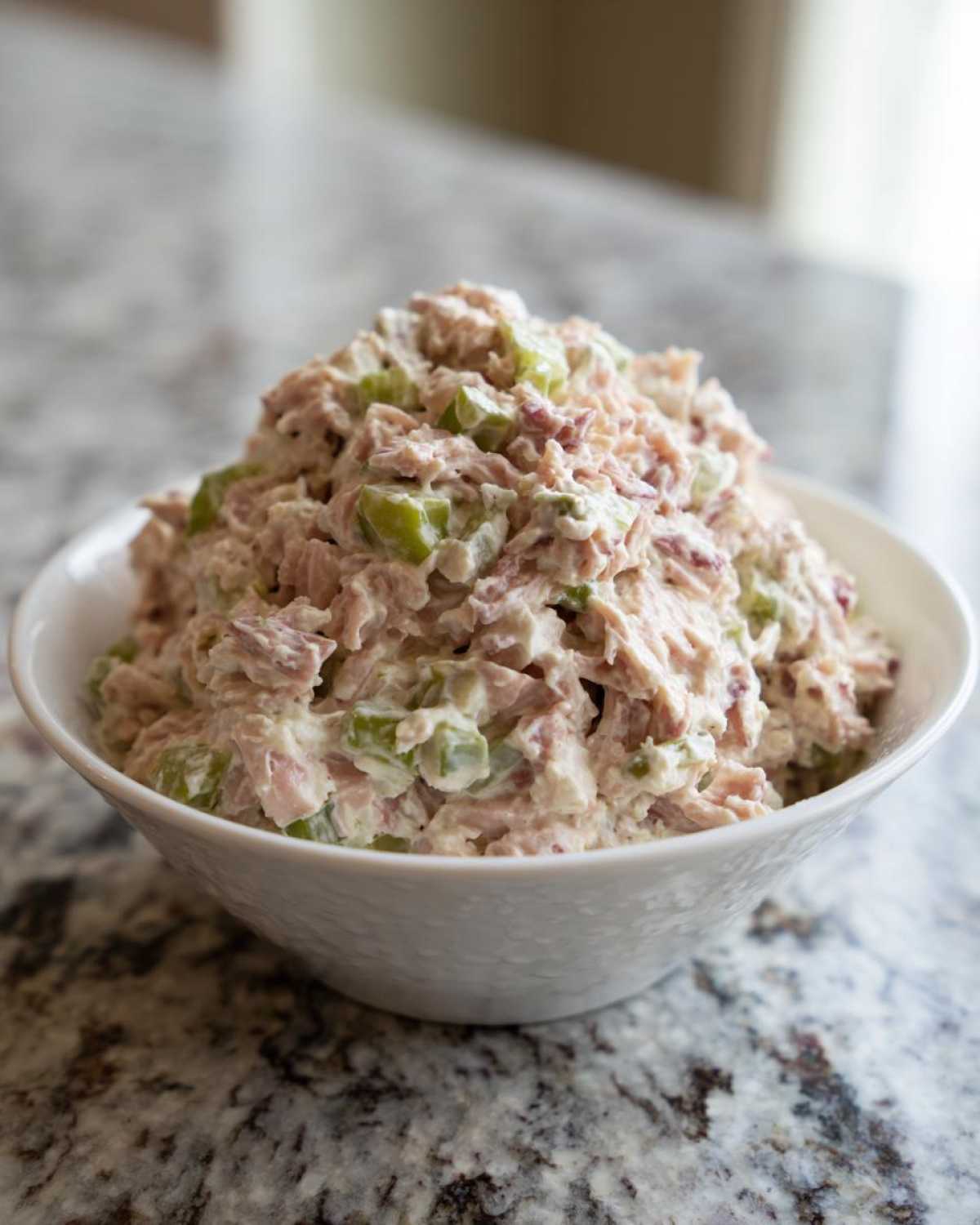 A white bowl filled with creamy ham salad recipe mixed with visible chunks of green celery, sitting on a granite countertop.