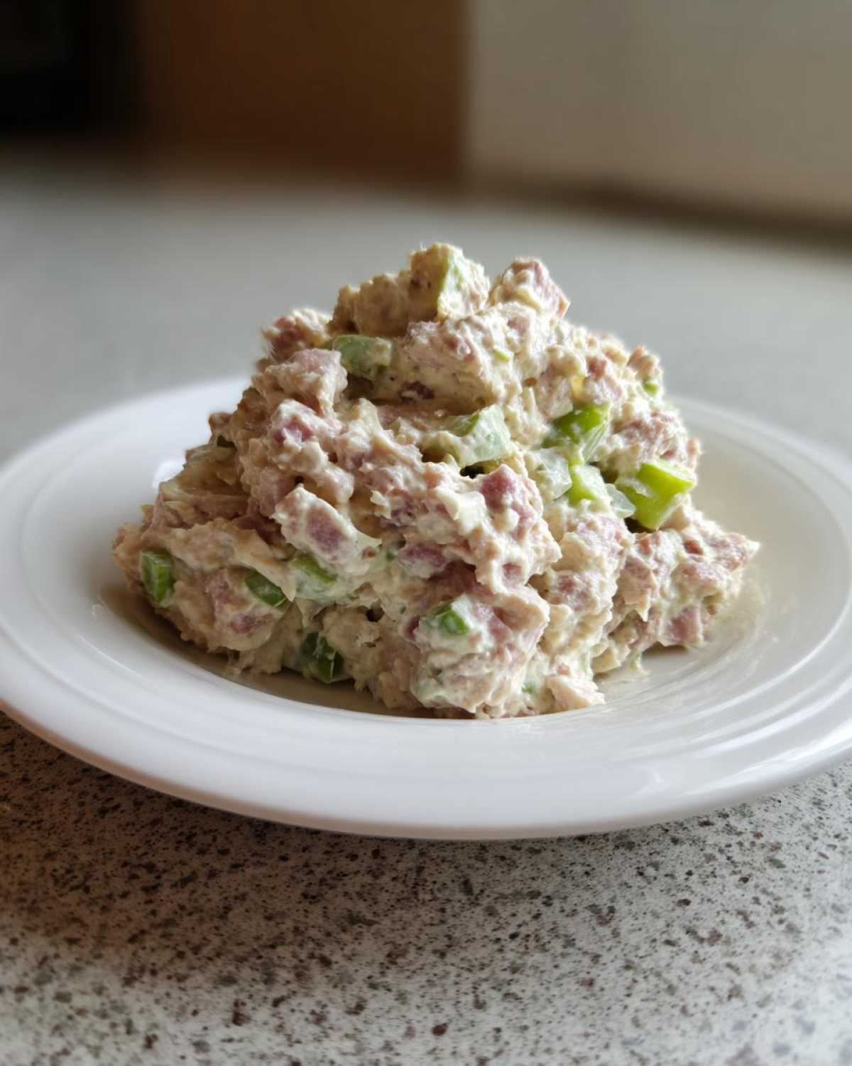 A mound of creamy ham salad recipe mixed with diced green celery served on a simple white plate.