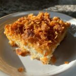 A square serving of creamy potato casserole topped generously with golden, crunchy cornflakes on a white plate.