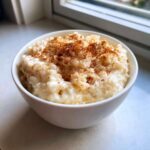 A close-up view of a creamy rice pudding recipe served in a white bowl, generously sprinkled with cinnamon.