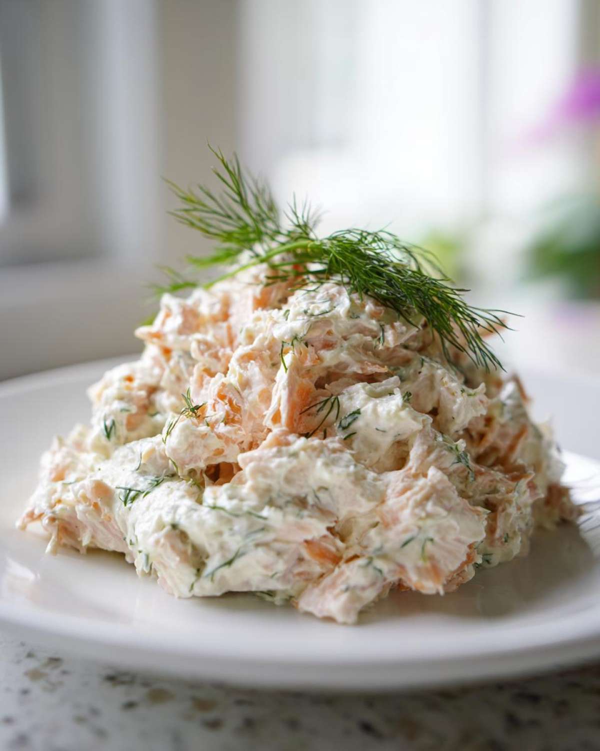 A mound of creamy salmon salad mixed with dill, garnished with a sprig of fresh dill, served on a white plate.