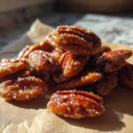 A pile of glistening, caramelized candied pecans sprinkled with coarse sugar resting on brown parchment paper.