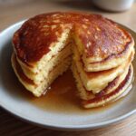 A stack of fluffy coconut flour pancakes with a piece cut out, drizzled generously with maple syrup.