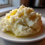 A generous serving of fluffy mashed potatoes topped with melted butter and cracked black pepper on a white plate.