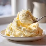 A spoonful of creamy, fluffy mashed potatoes seasoned with black pepper being lifted from a serving on a white plate.