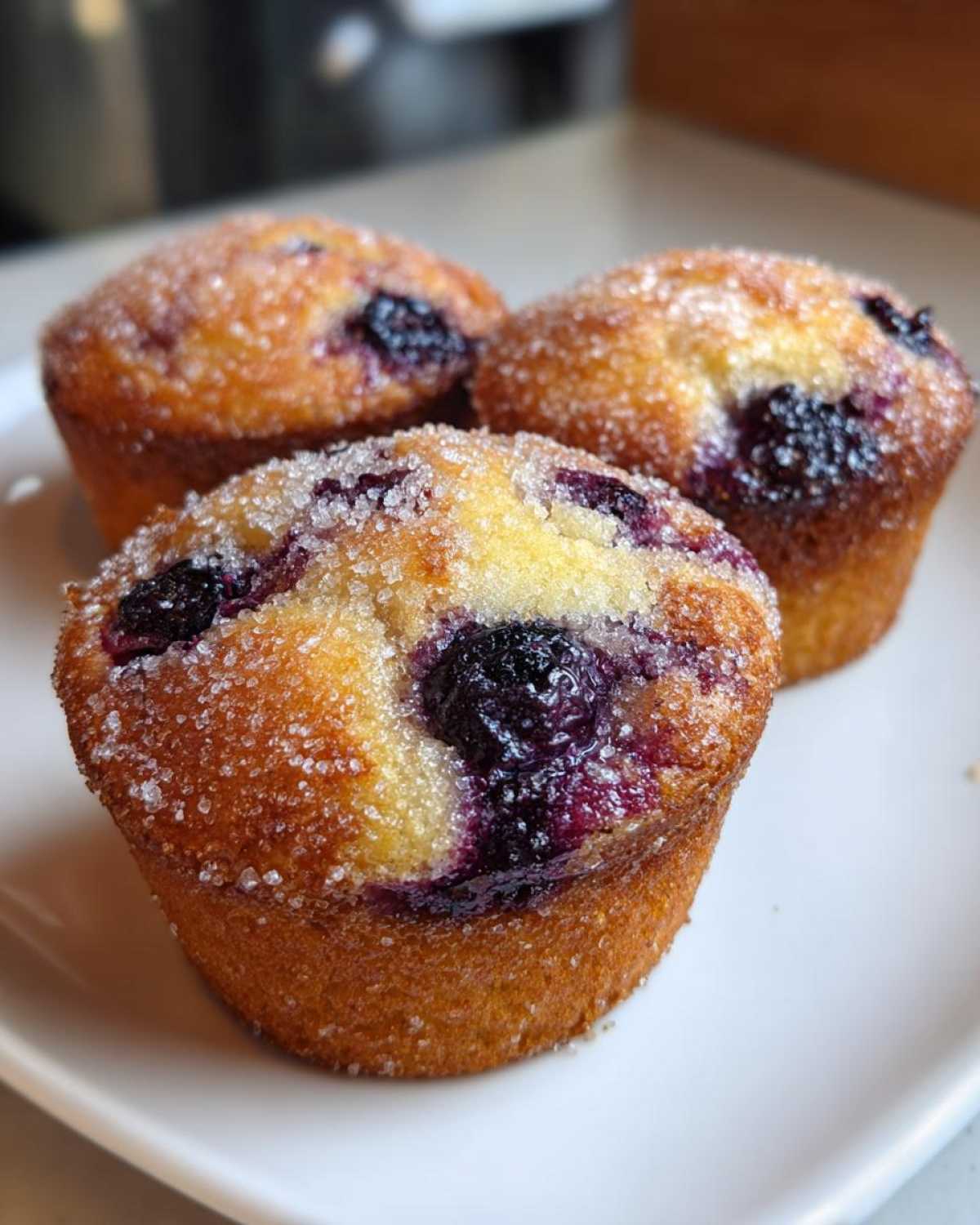Three golden Gluten-Free Blueberry Lemon Muffin Tops topped with coarse sugar crystals, sitting on a white plate.