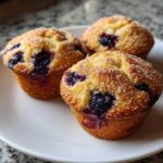 Three golden Gluten-Free Blueberry Lemon Muffin Tops sprinkled generously with coarse sugar, sitting on a white plate.