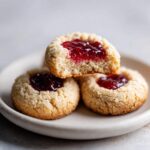 Three Gluten-Free Raspberry Almond Thumbprint Cookies stacked on a plate, one bitten open showing the jam center.