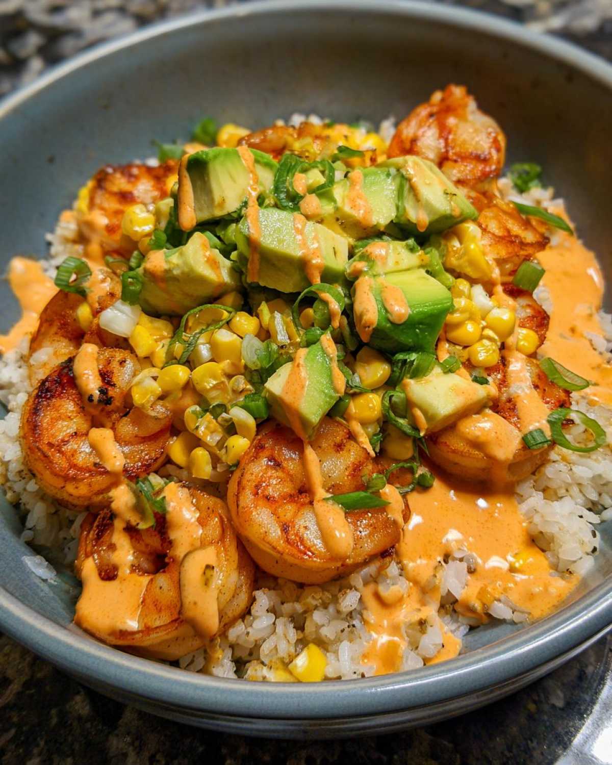 Close-up of a gourmet shrimp bowl featuring seasoned shrimp, rice, corn, avocado chunks, and a creamy orange sauce.