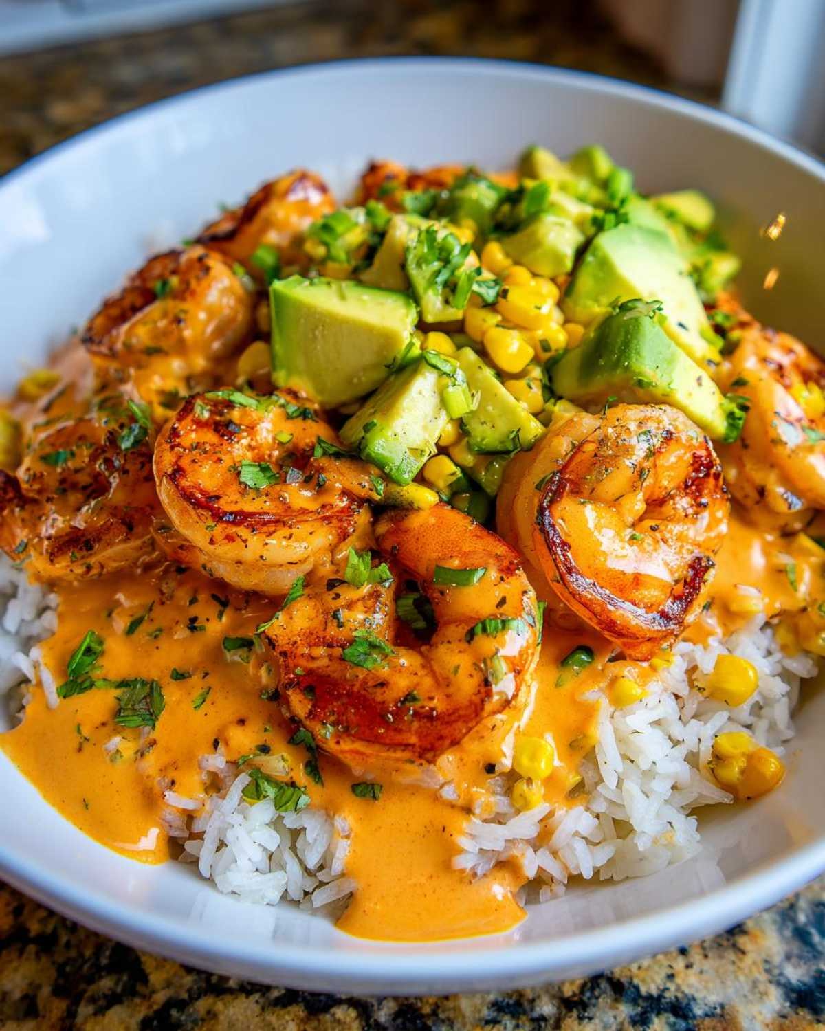 Close-up of a gourmet shrimp bowl featuring seasoned shrimp, white rice, corn, avocado chunks, and a creamy orange sauce.