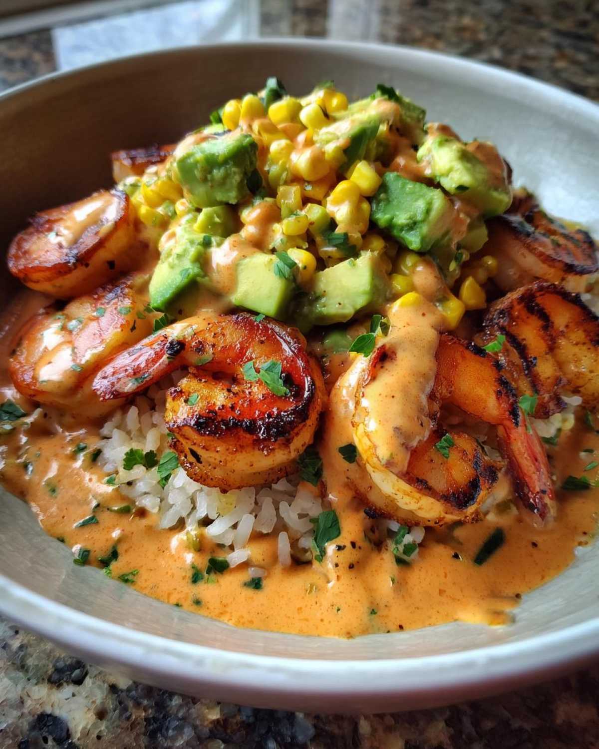 Close-up of a gourmet shrimp bowl featuring grilled shrimp, rice, creamy orange sauce, avocado chunks, and corn.