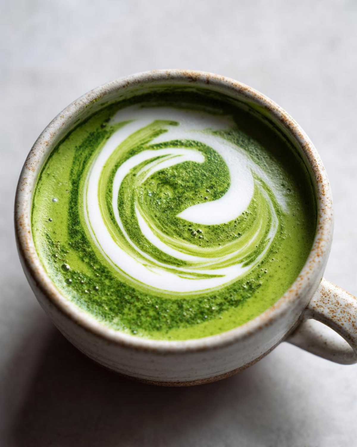 Close-up of a vibrant green drink with white foam art swirled on top, served in a rustic ceramic mug.