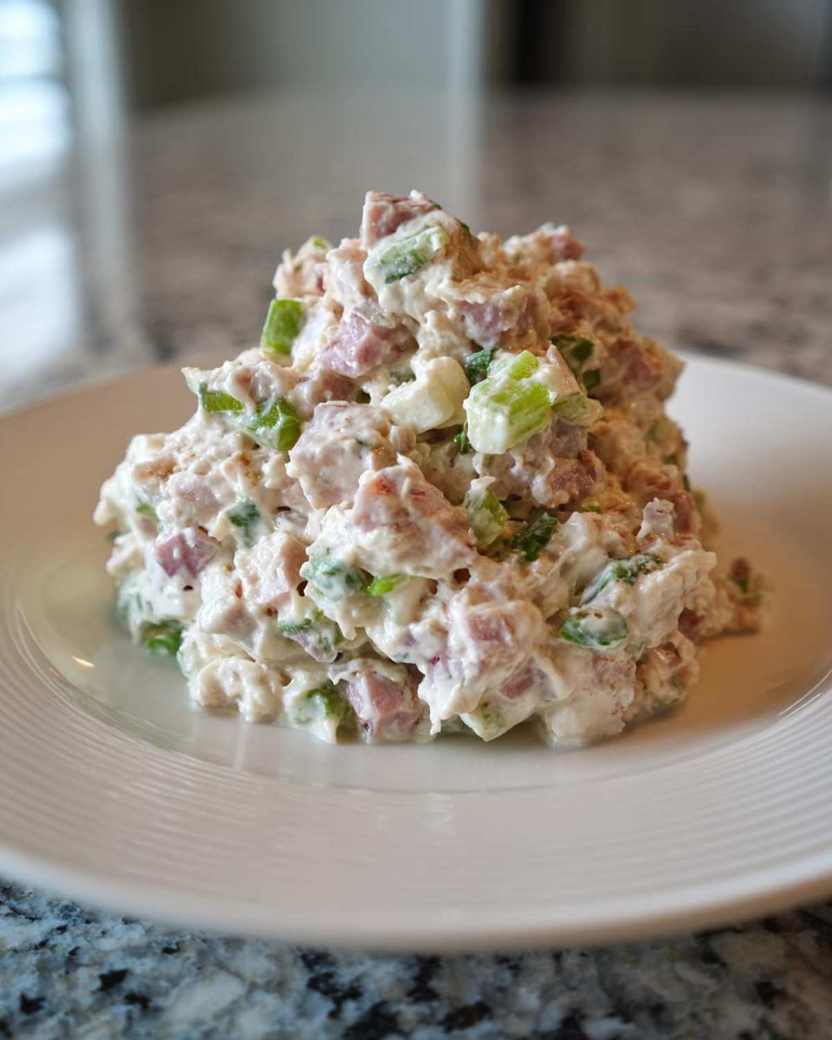 A mound of creamy, chunky ham salad recipe mixed with green celery pieces served on a white plate.
