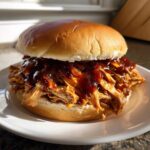 A close-up of a fully loaded Instant Pot BBQ Pulled Chicken Sandwich on a white plate, dripping with sauce.