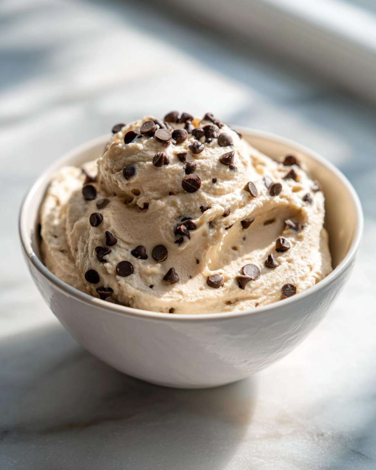 A close-up of creamy Keto Chocolate Chip Cookie Dough Dip topped generously with mini chocolate chips in a white bowl.