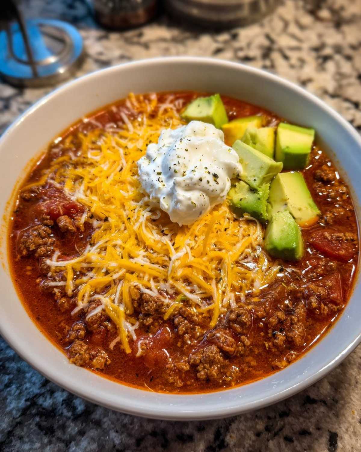 A close-up of a bowl of rich Keto Taco Soup with Ground Beef, topped with shredded cheese, sour cream, and diced avocado.