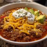A close-up of a bowl of Keto Taco Soup with Ground Beef, topped with shredded cheese, sour cream, and avocado slices.