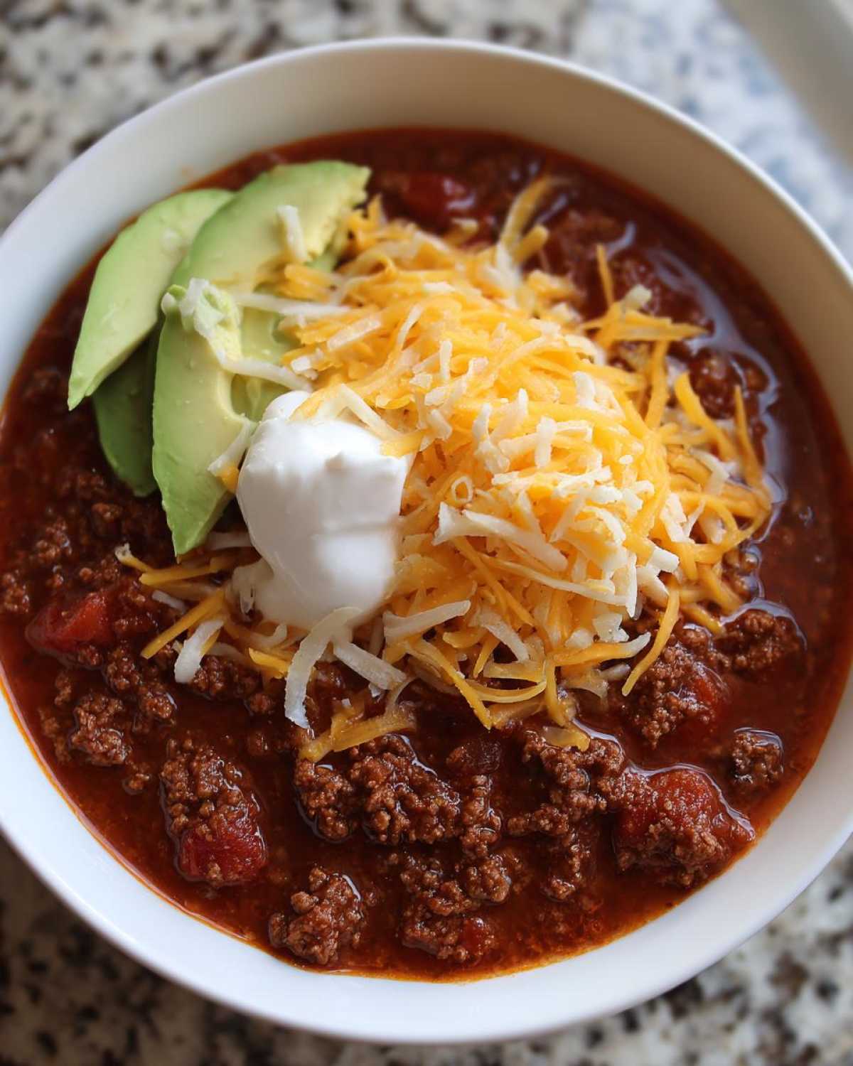 Close-up of a bowl of rich Keto Taco Soup with Ground Beef, topped with shredded cheese, sour cream, and avocado slices.