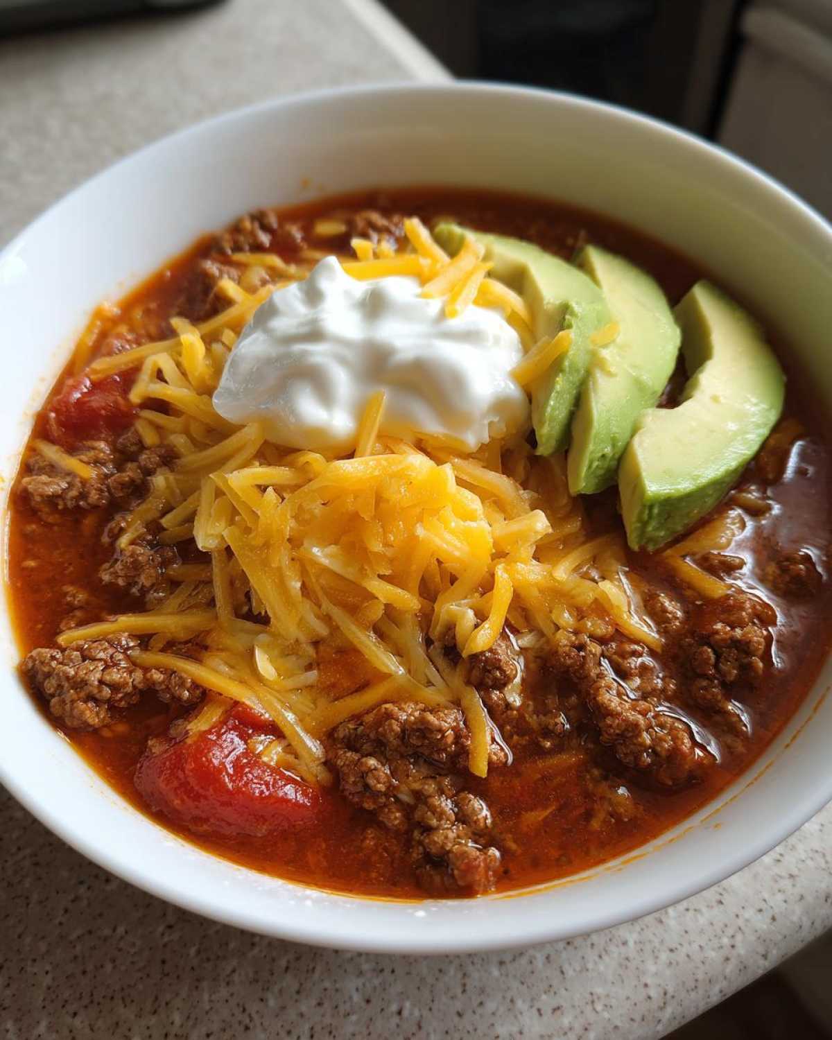 A close-up of a bowl of Keto Taco Soup with Ground Beef, topped with shredded cheese, sour cream, and avocado slices.