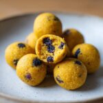 A stack of bright yellow No-Bake Lemon Blueberry Energy Bites, with one bitten in half showing the blueberry pieces inside.