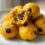 Close-up of vibrant yellow No-Bake Lemon Blueberry Energy Bites, one is broken open showing dried blueberries inside.