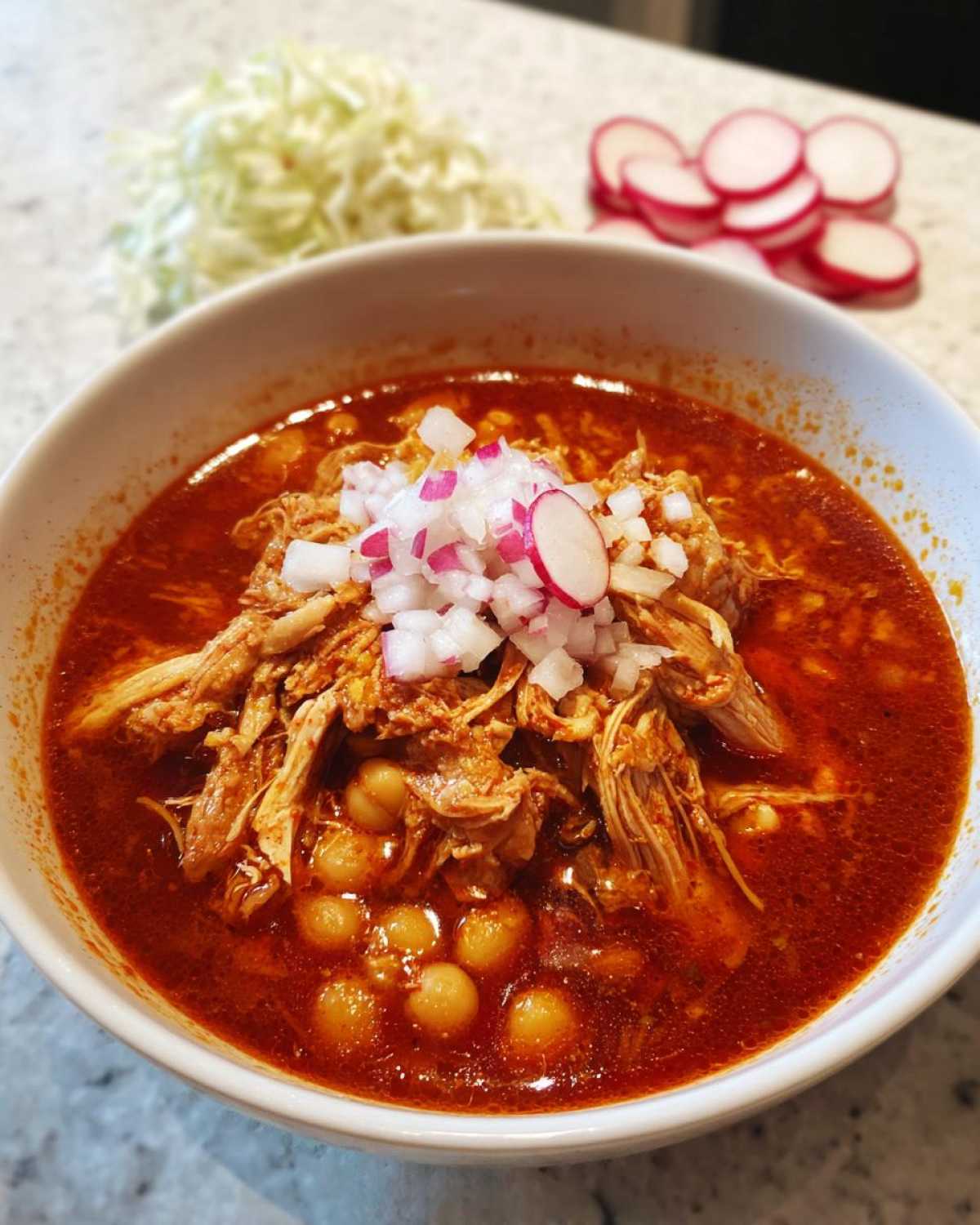 A close-up of a bowl of rich red chicken pozole topped with shredded chicken, hominy, diced onion, and radish.