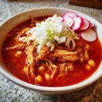 A vibrant bowl of red chicken pozole topped with shredded chicken, cabbage, onion, and sliced radishes.