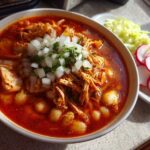 A vibrant bowl of red chicken pozole, filled with shredded chicken and hominy, topped with diced white onion and cilantro.