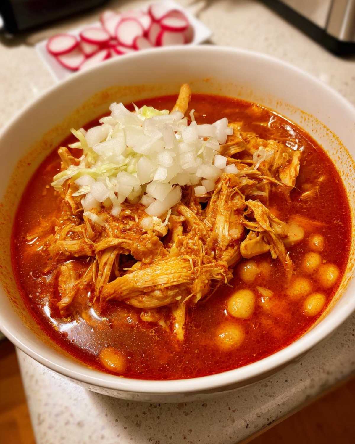 Close-up of a bowl of rich red chicken pozole topped with shredded chicken, onions, and hominy.