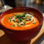 Close-up of a vibrant red tomato bisque in a rustic bowl, swirled with cream and topped with fresh basil.