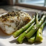 A perfectly cooked fillet of Sheet Pan Lemon Herb Cod next to bright green asparagus spears on a white plate.