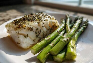 A perfectly cooked fillet of Sheet Pan Lemon Herb Cod next to bright green asparagus spears on a white plate.