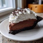 A decadent slice of chocolate mousse pie featuring a dark crust, rich chocolate filling, and a thick layer of whipped cream topped with chocolate shavings.