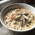 A close-up of a white bowl filled with Slow Cooker Creamy Chicken and Wild Rice Soup, showing shredded chicken, wild rice, carrots, and mushrooms.