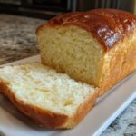 A golden-brown loaf of soft condensed milk bread, with one thick slice cut and resting beside it on a white platter.